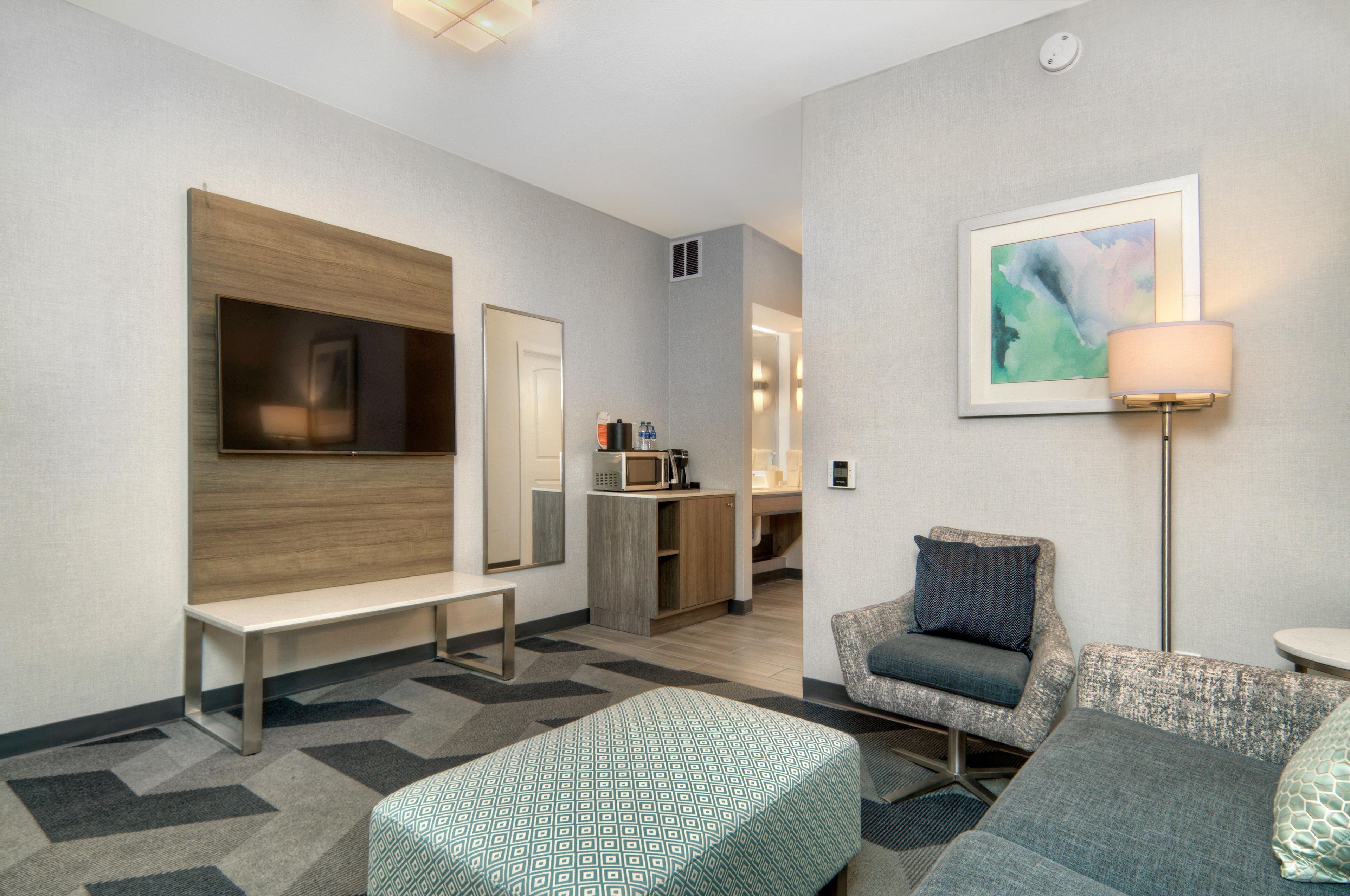 A hotel room with a TV, a bench, a sofa, a coffee machine, and a painting on the wall.
