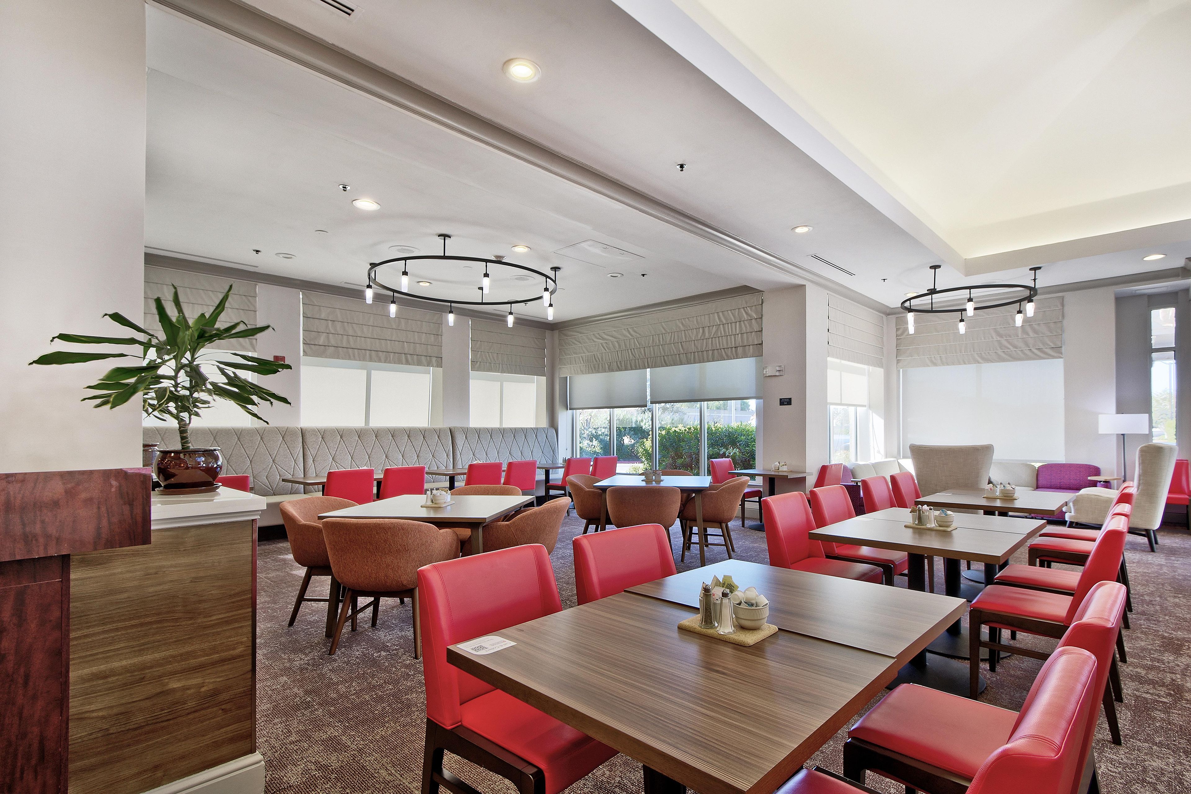 A hotel dining area with multiple tables and chairs, large windows, and a potted plant.