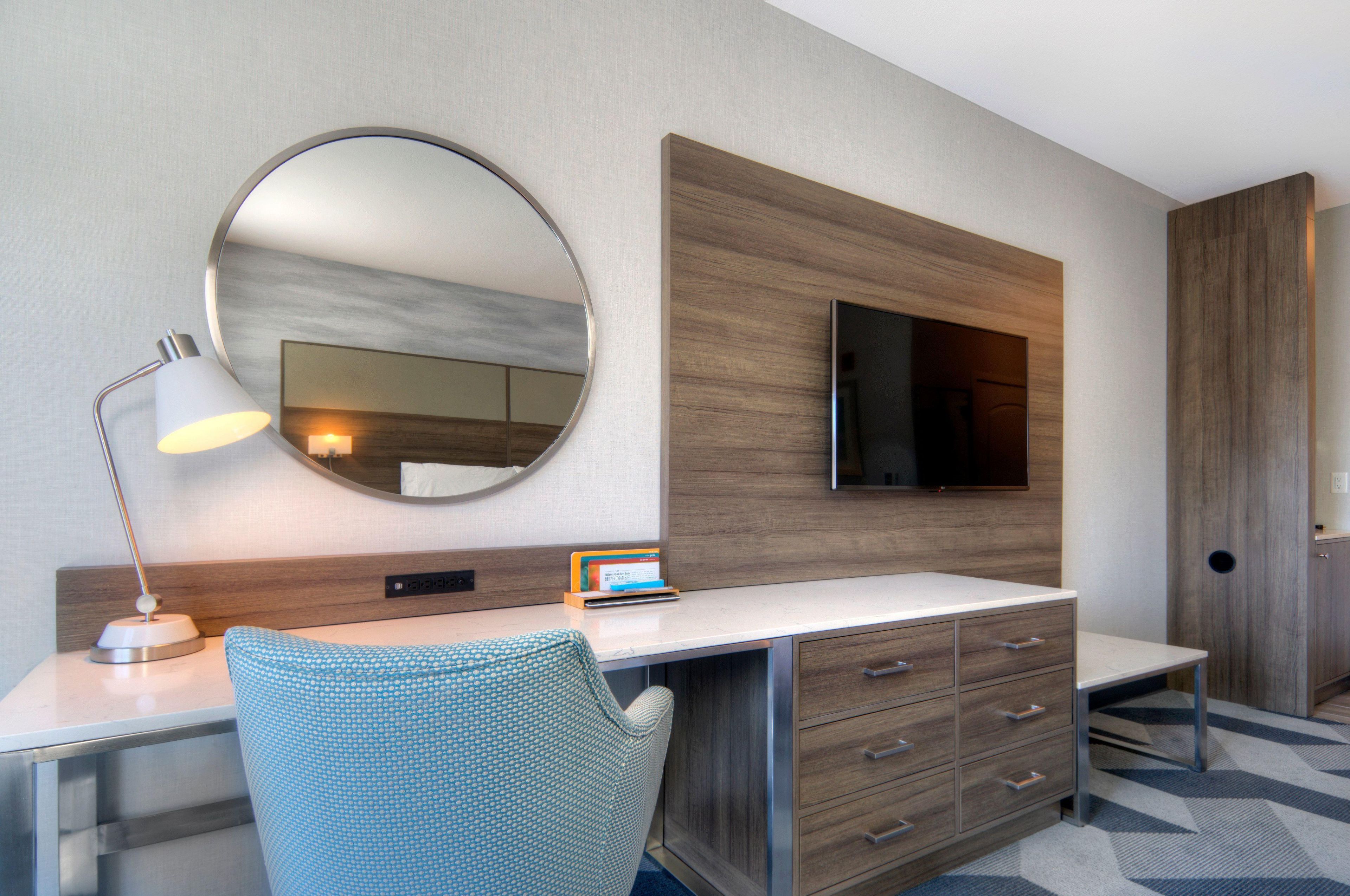 A modern hotel room with a desk, a round mirror, a flat-screen TV, and a seating area with a patterned chair.