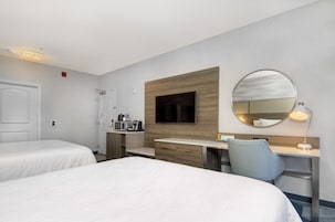 A hotel room with two beds, a desk with a chair, a TV, a mirror, and a small kitchenette.