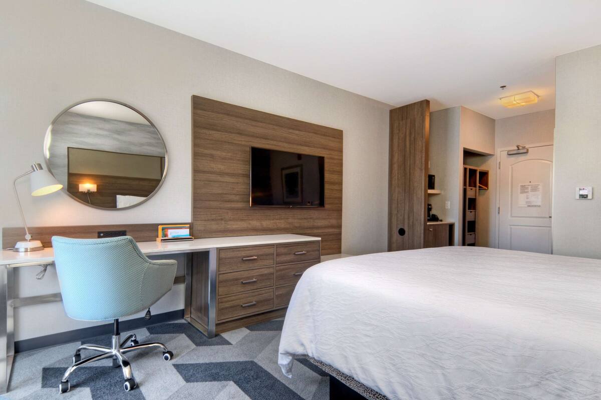 Habitación de hotel con cama, escritorio con silla, televisión montada en la pared y espejo.