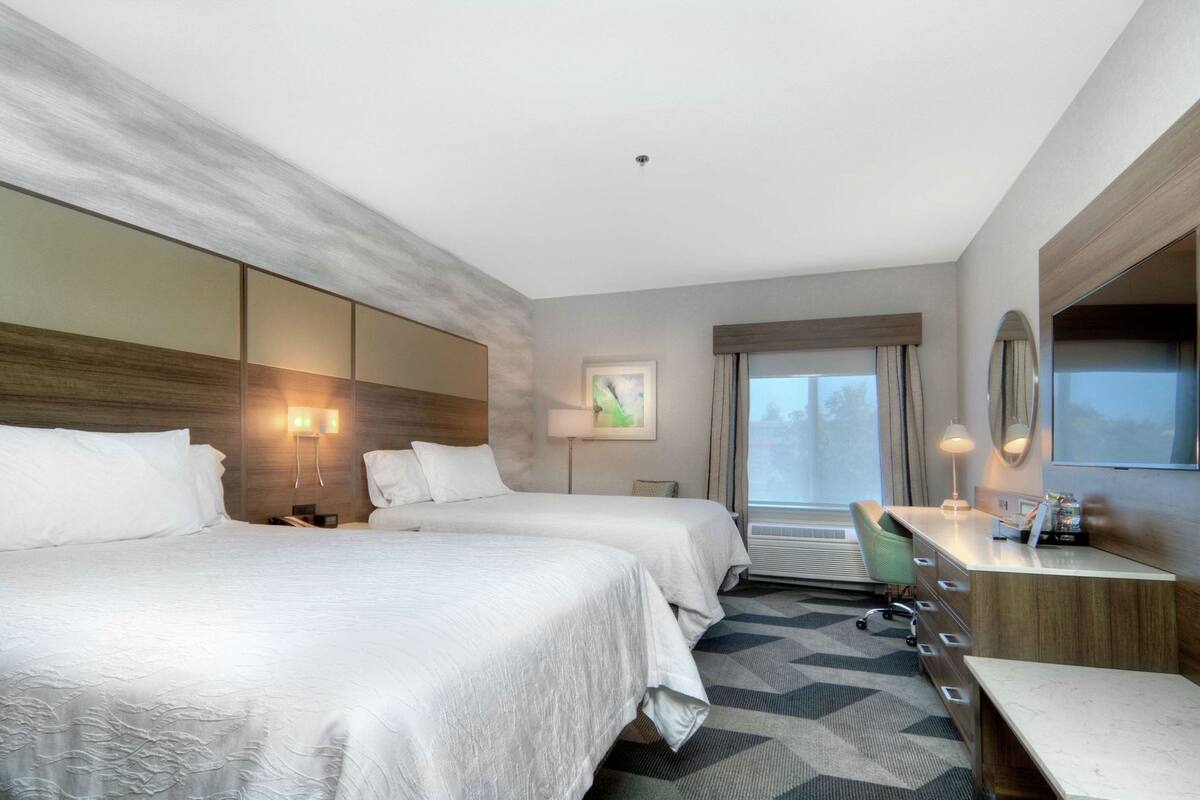 Habitación de hotel con dos camas, un escritorio, una silla, un espejo grande y una ventana con cortinas.