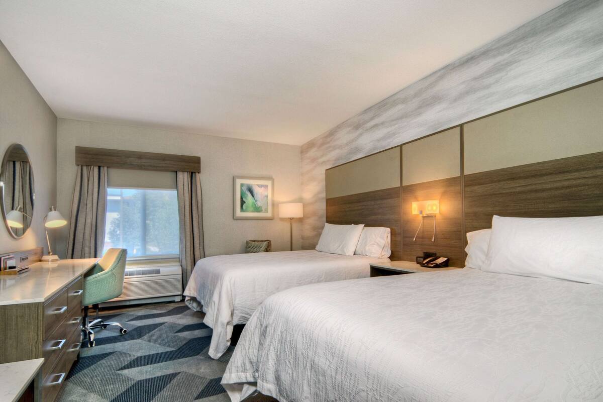Habitación de hotel con dos camas, un escritorio con silla, una lámpara y un cuadro en la pared.