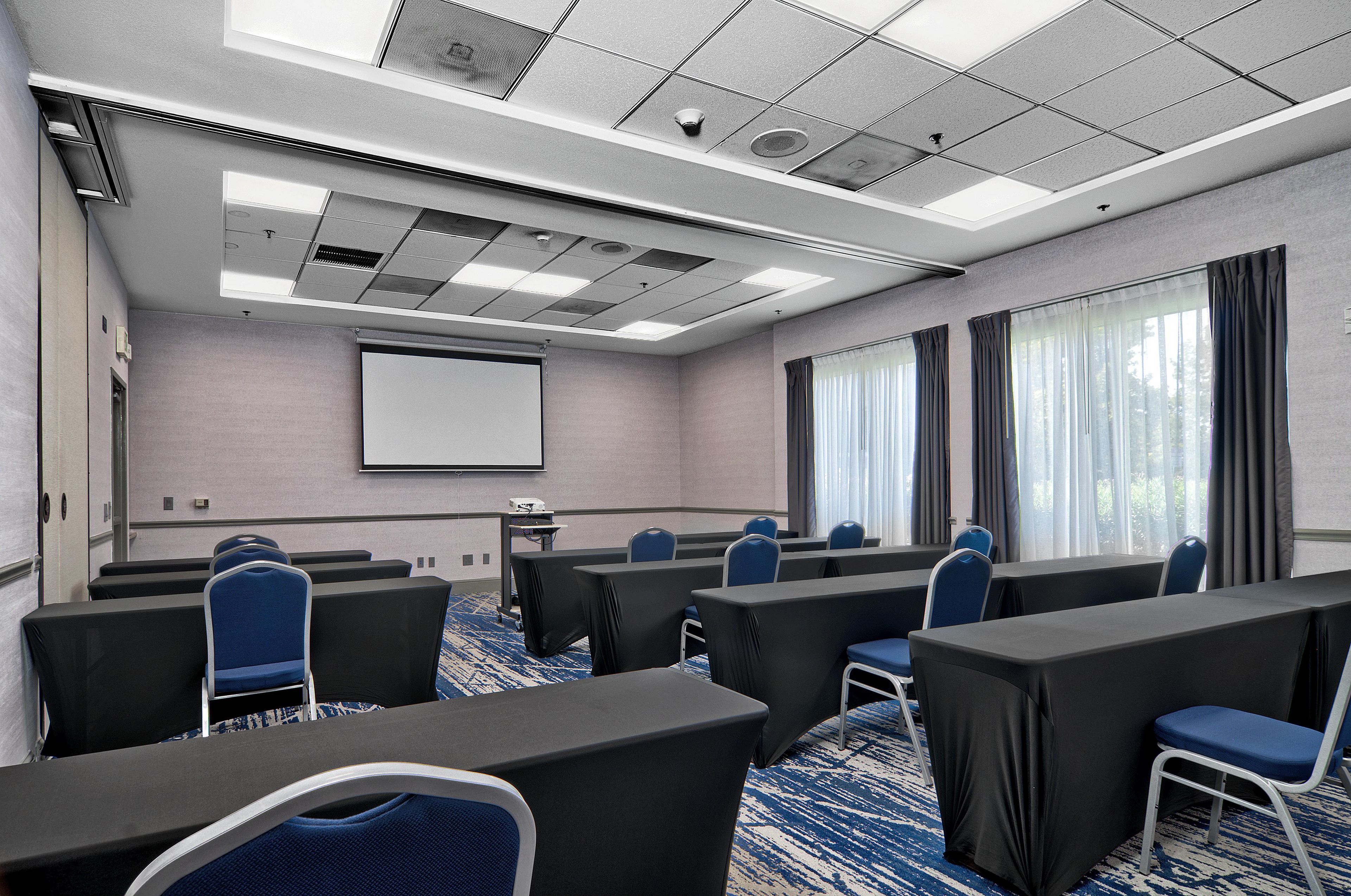 A conference room with a projector screen, rows of tables, chairs, and a carpeted floor.