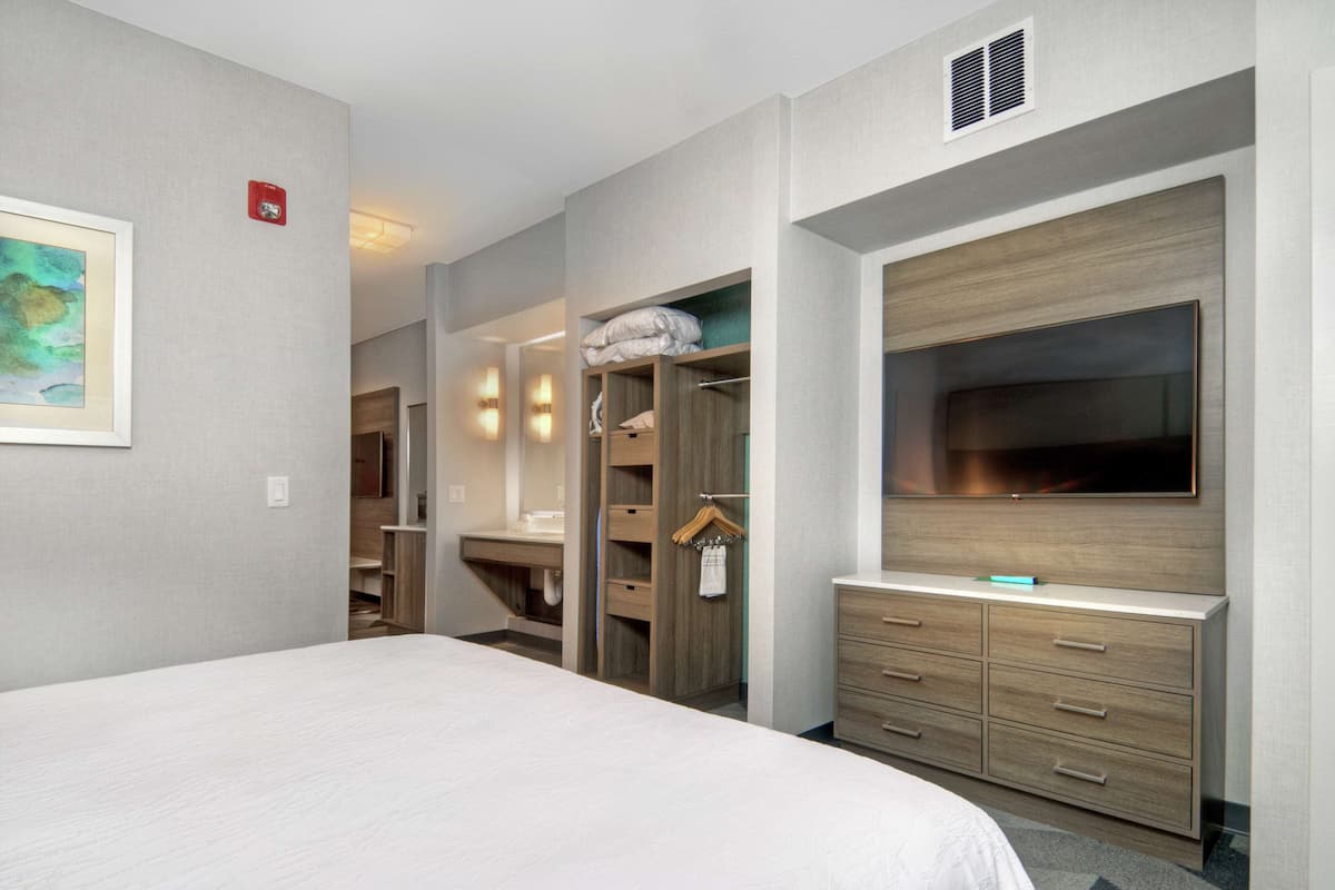Habitación de hotel con una cama grande, televisión montada en la pared, vestidor y baño con ducha e inodoro.