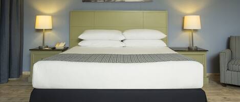 A neatly made bed with a headboard, two bedside tables with lamps, a chair, and a framed picture on the wall.
