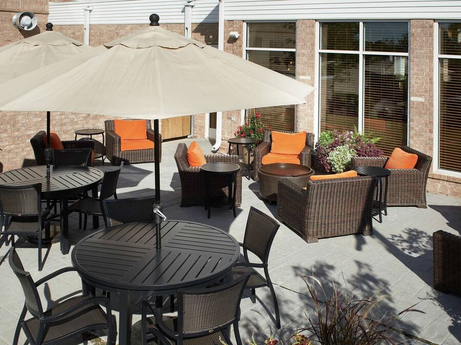 An outdoor patio with round tables and wicker chairs, some with orange cushions, and large umbrellas providing shade.