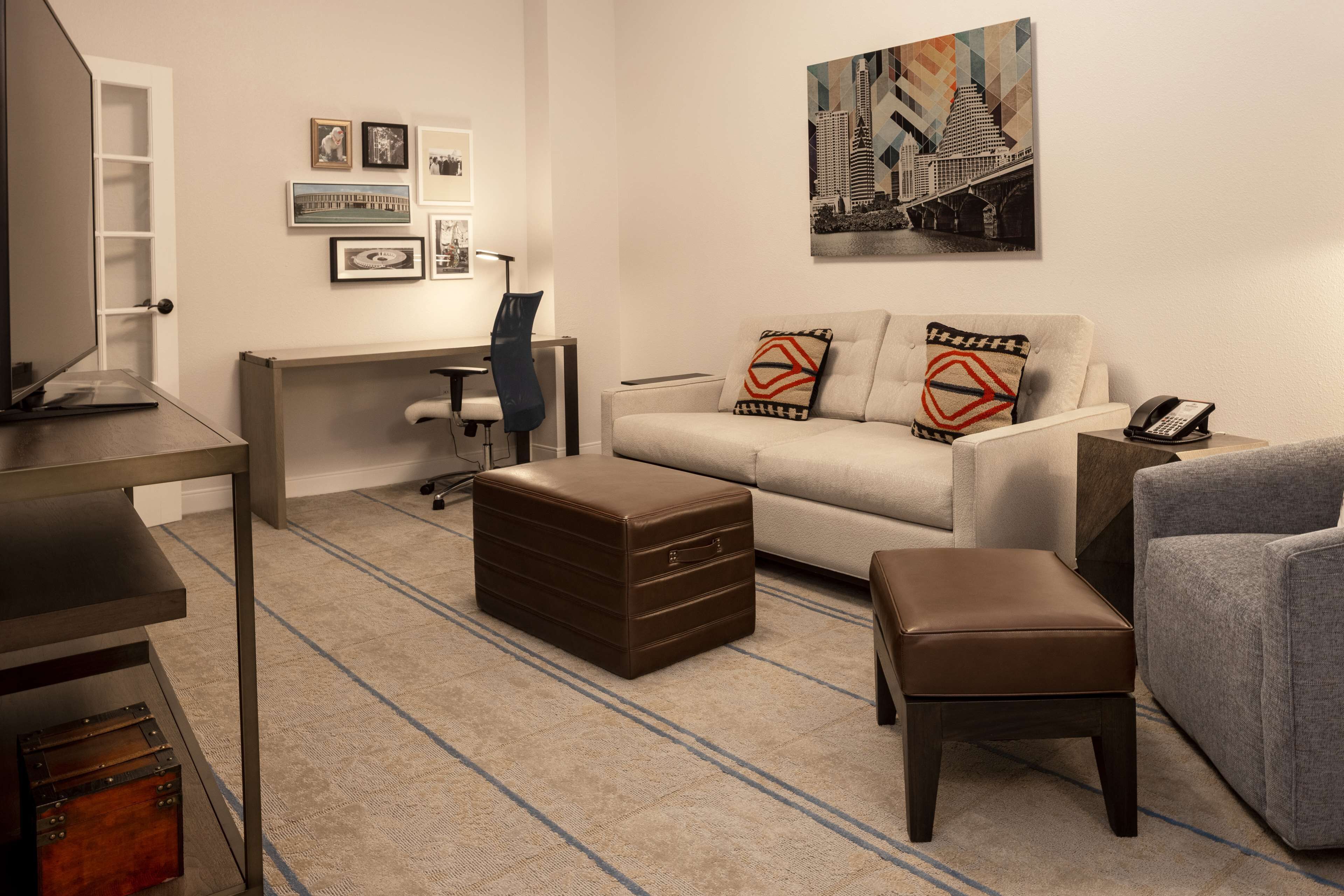 Una sala de estar moderna con un sofá, un reposapiés y un escritorio con una silla. Hay cuadros enmarcados en la pared y un cuadro sobre el sofá.