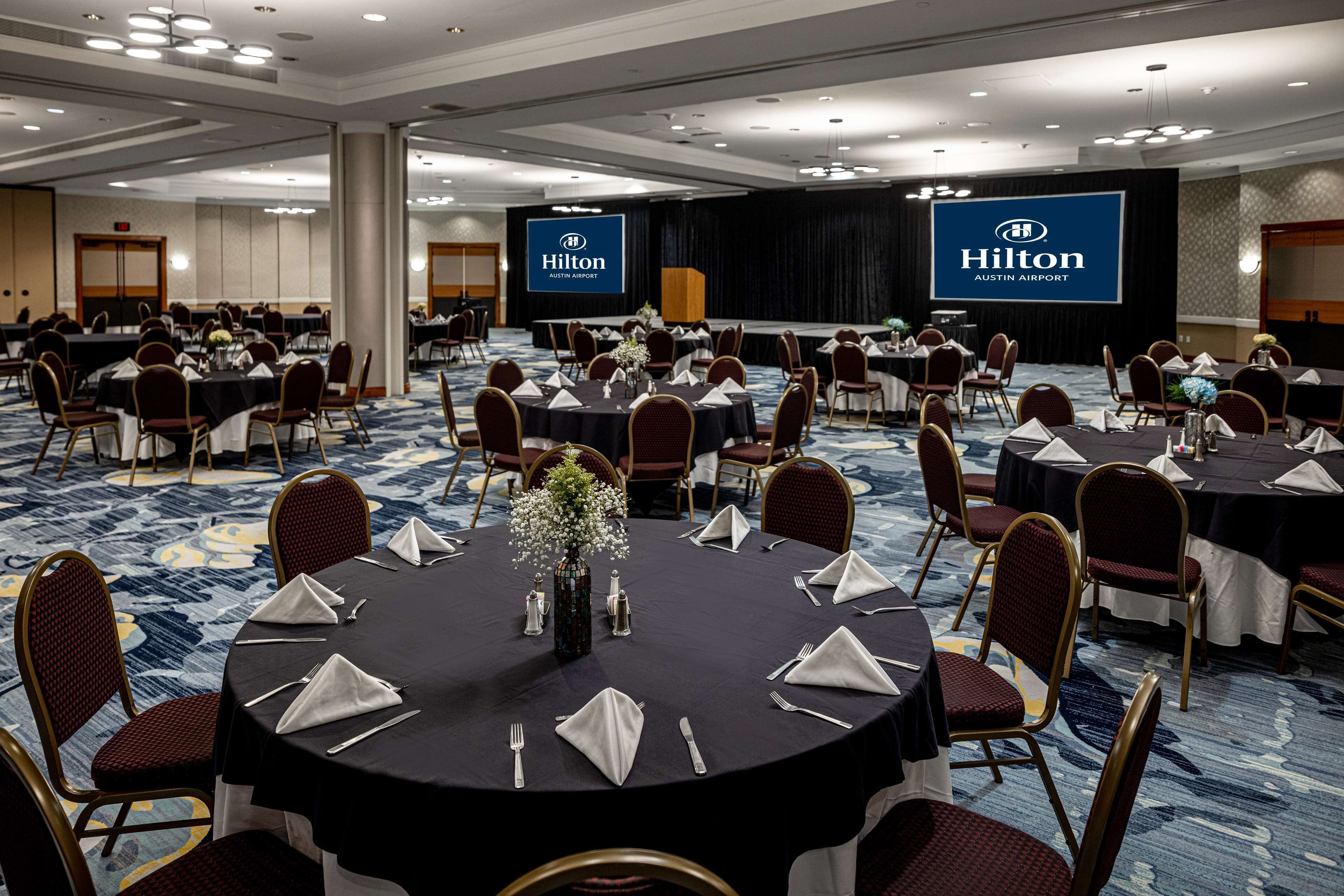 A large conference room with multiple tables set for a meeting, featuring a Hilton logo and a floral centerpiece.