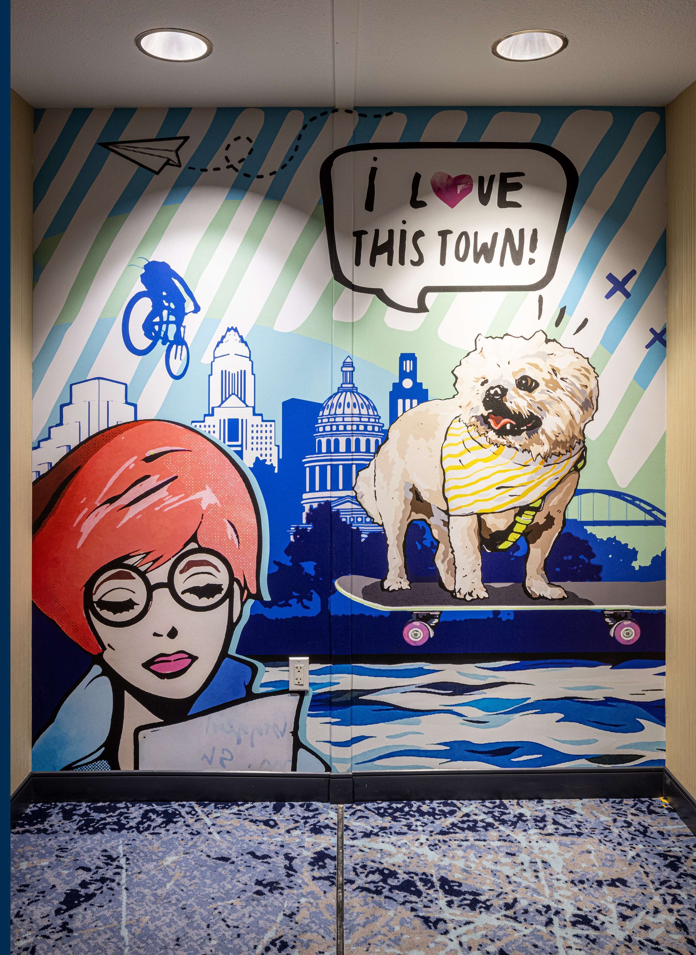 Un mural con un perro y una persona en un monopatín, una burbuja de diálogo que dice "¡ME ENCANTA ESTA CIUDAD!", y un paisaje urbano al fondo.