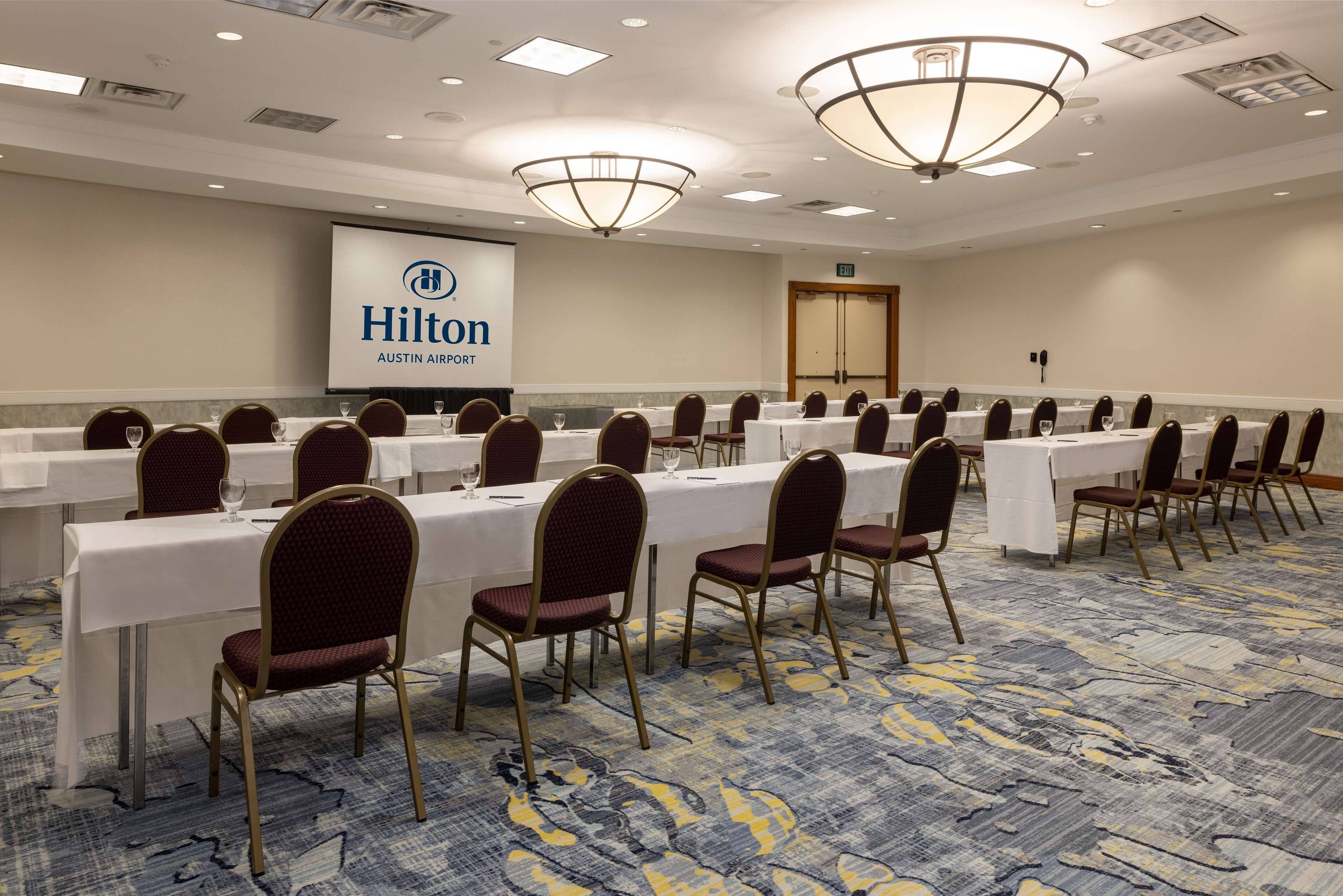A conference room with long tables, chairs, and a large screen displaying the Hilton logo.