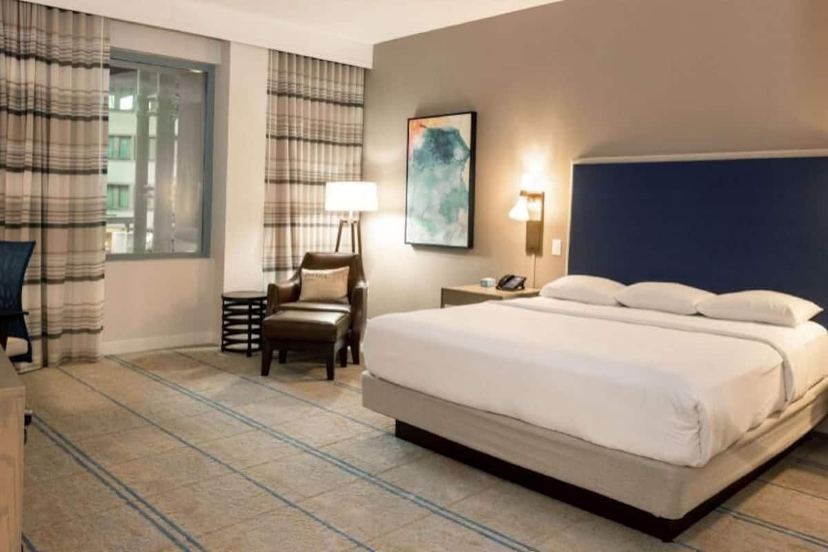 A hotel room with a large bed, a desk, a chair, a nightstand, and a window with curtains.