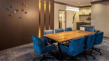A conference room with a wooden table and blue chairs, a wooden partition with hanging figurines, and a kitchen area with a sink and countertop.