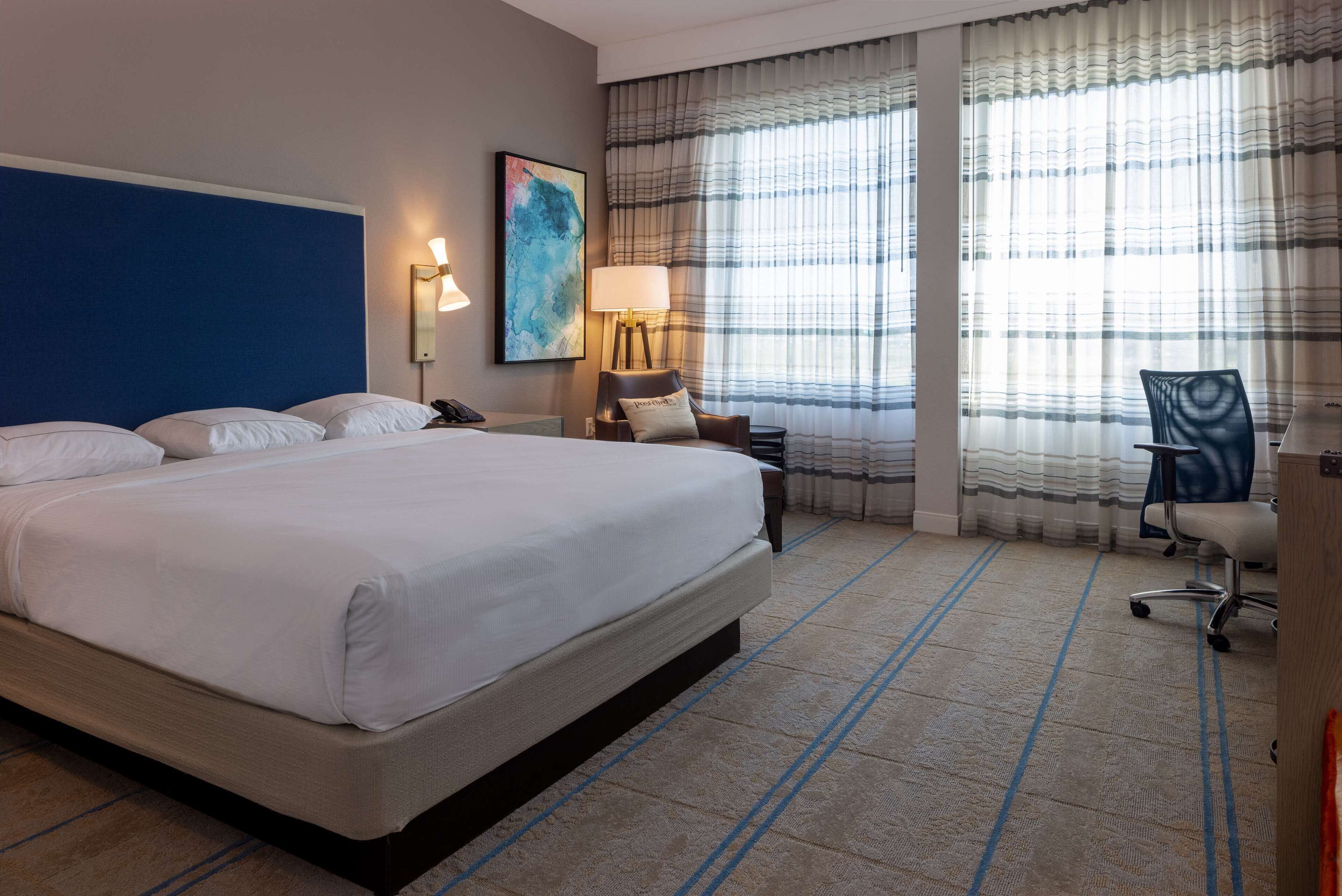 Une chambre d’hôtel avec un grand lit, un bureau, une chaise, une lampe et une fenêtre avec des rideaux.