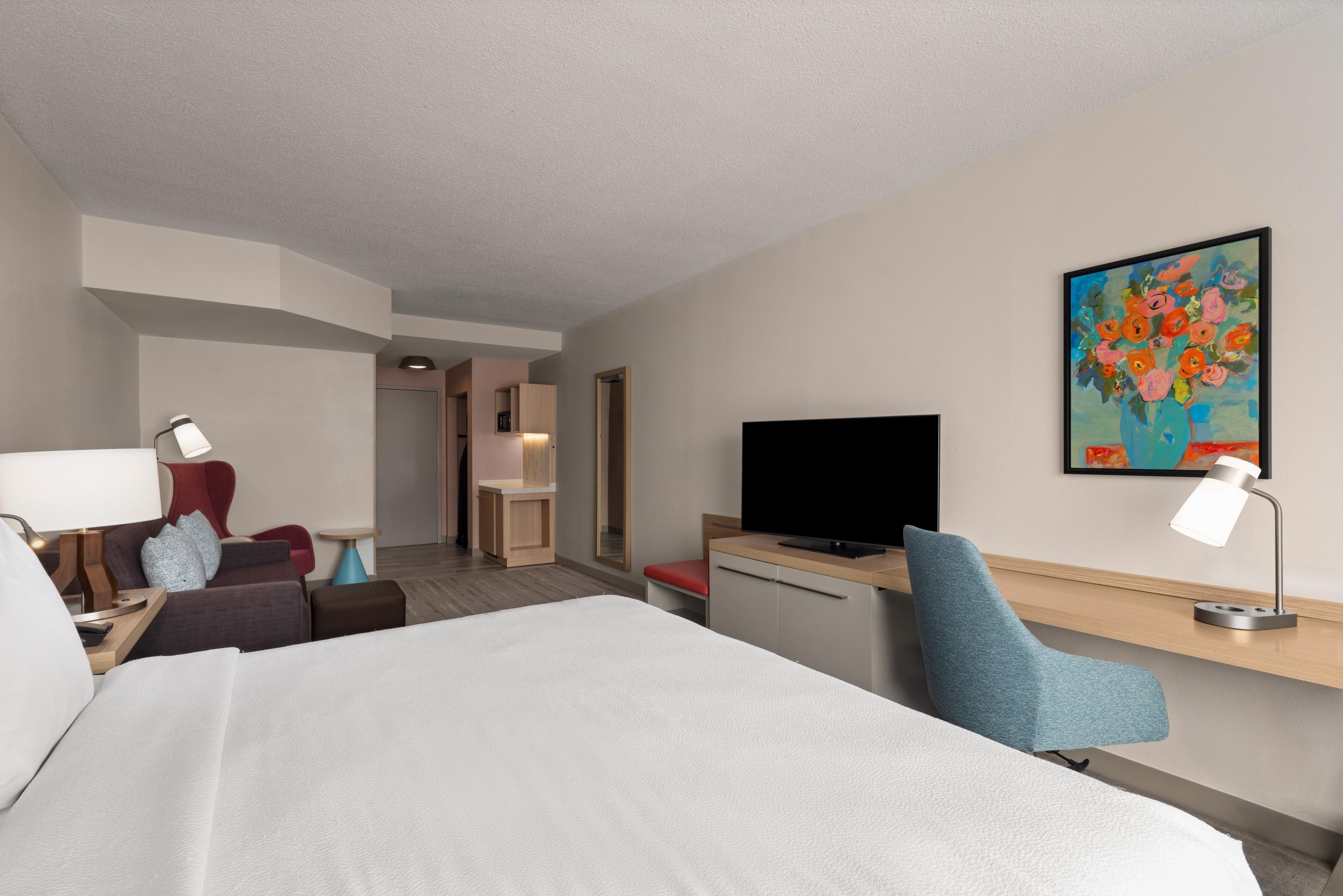 A hotel room with a bed, desk, chair, TV, and artwork on the wall.