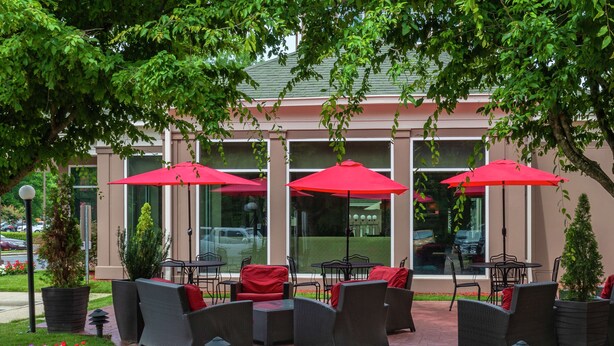 An outdoor seating area with wicker furniture and red umbrellas, surrounded by flower beds and a building with large windows. - Hilton Garden Inn Charlotte Pineville (Pineville)