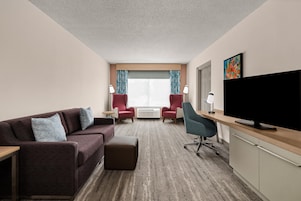 A hotel room with a sofa, two armchairs, a desk with a flat-screen TV, and a large window with curtains.