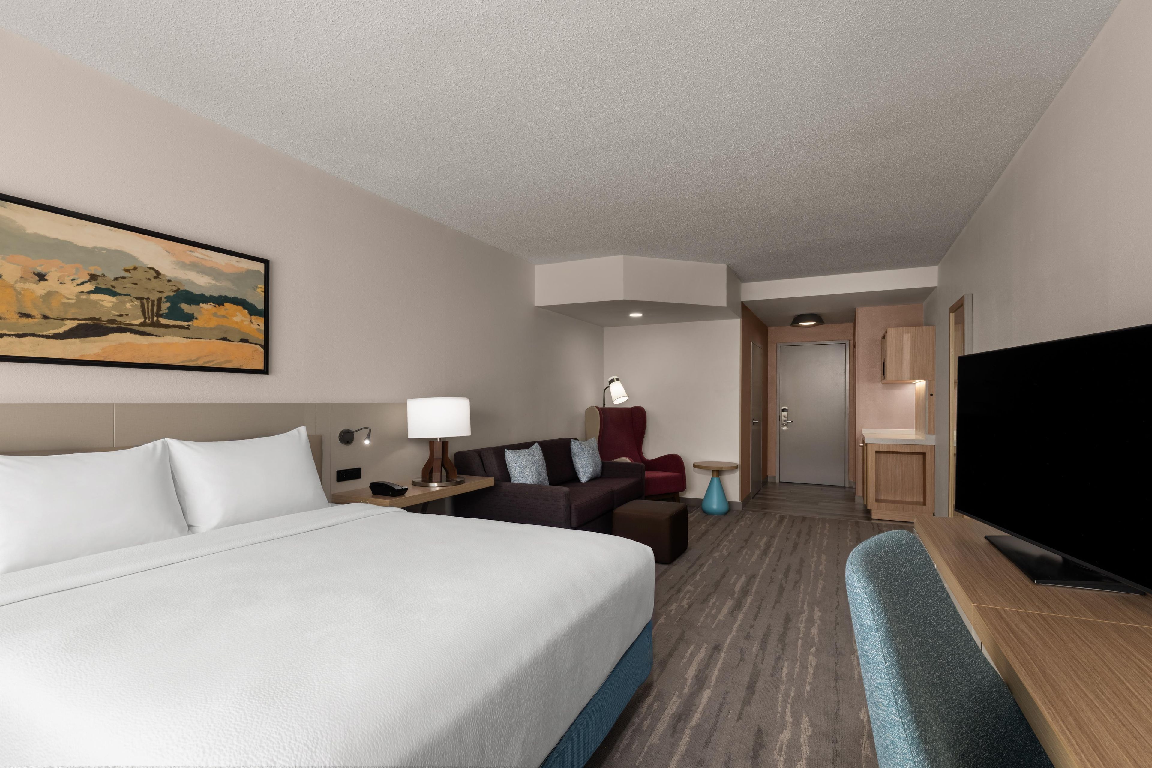 A hotel room with a bed, a desk, a TV, a sofa, and a painting on the wall.