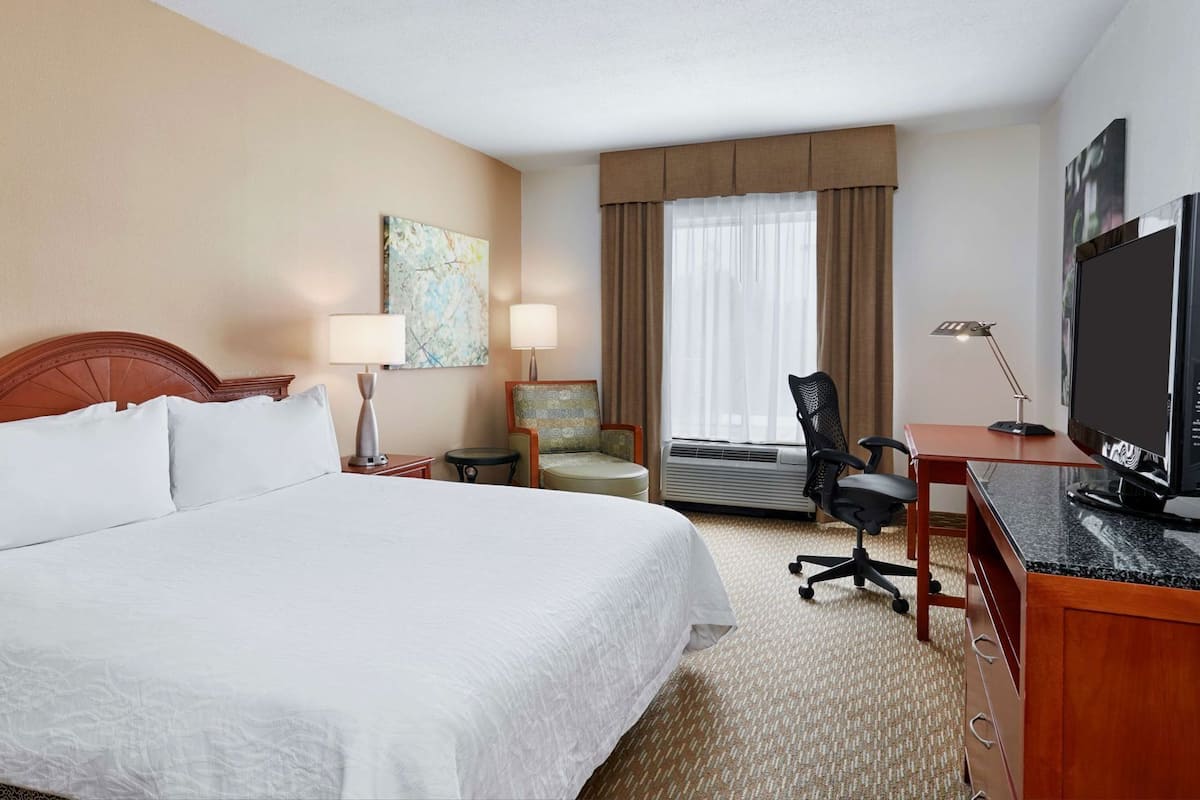 A hotel room with a large bed, a desk with a TV, a chair, a lamp, and a painting on the wall.