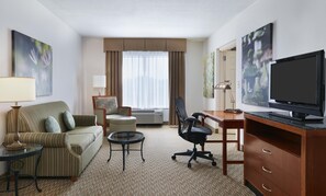 Ein Hotelzimmer mit einer Couch, einem Schreibtisch mit Computer, einem Fernseher und Kunstwerken an den Wänden.