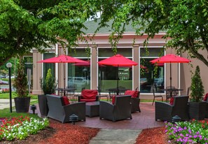An outdoor seating area with wicker furniture and red umbrellas, in front of a building with large windows. - Hilton Garden Inn Charlotte Pineville (Pineville)