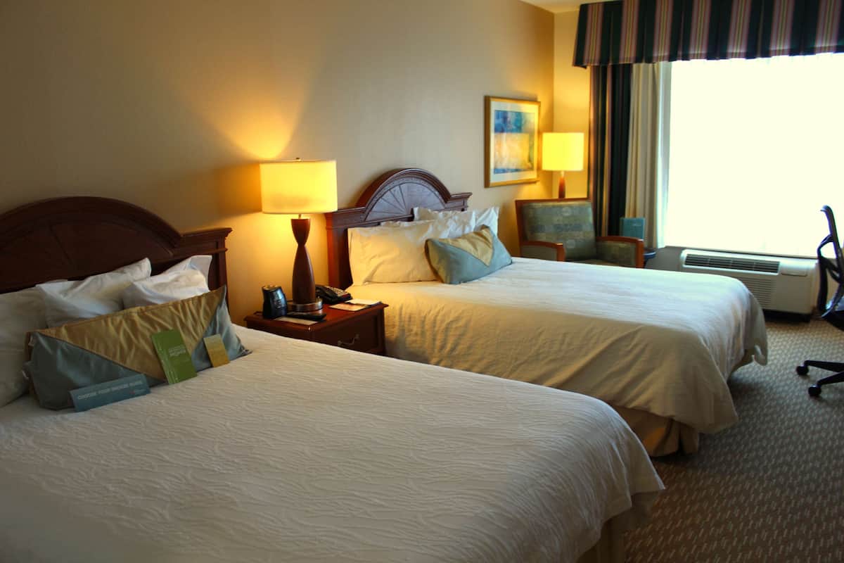 A hotel room with two beds, bedside lamps, a nightstand, a chair, and a window with curtains.