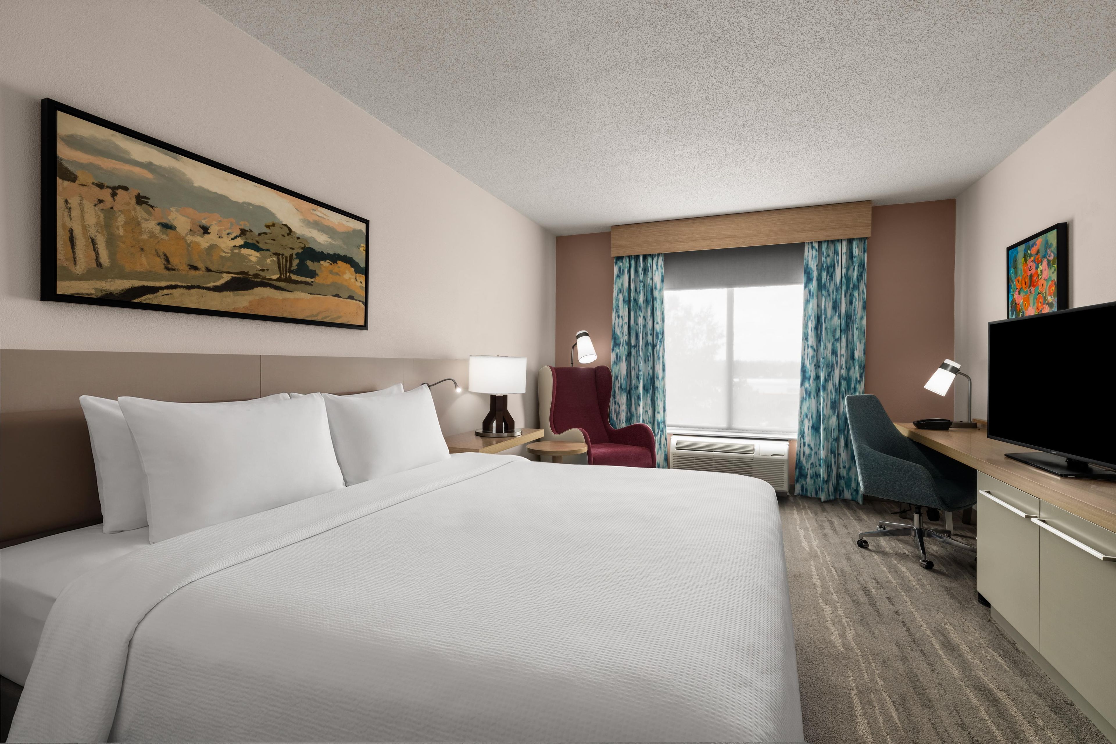 A hotel room with a large bed, a desk with a computer, a chair, a TV, and a painting on the wall.