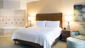 A hotel room with a large bed, two bedside lamps, a nightstand, a chair, a painting, and a bathroom with a bathtub and sink. - Hilton Garden Inn Charlotte Pineville (Pineville)