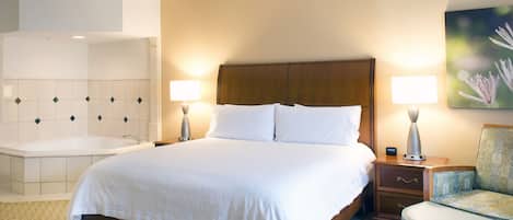 Una camera d'albergo con un letto grande, due lampade da comodino, un tavolino, una sedia, un quadro e un bagno con una vasca e un lavandino.