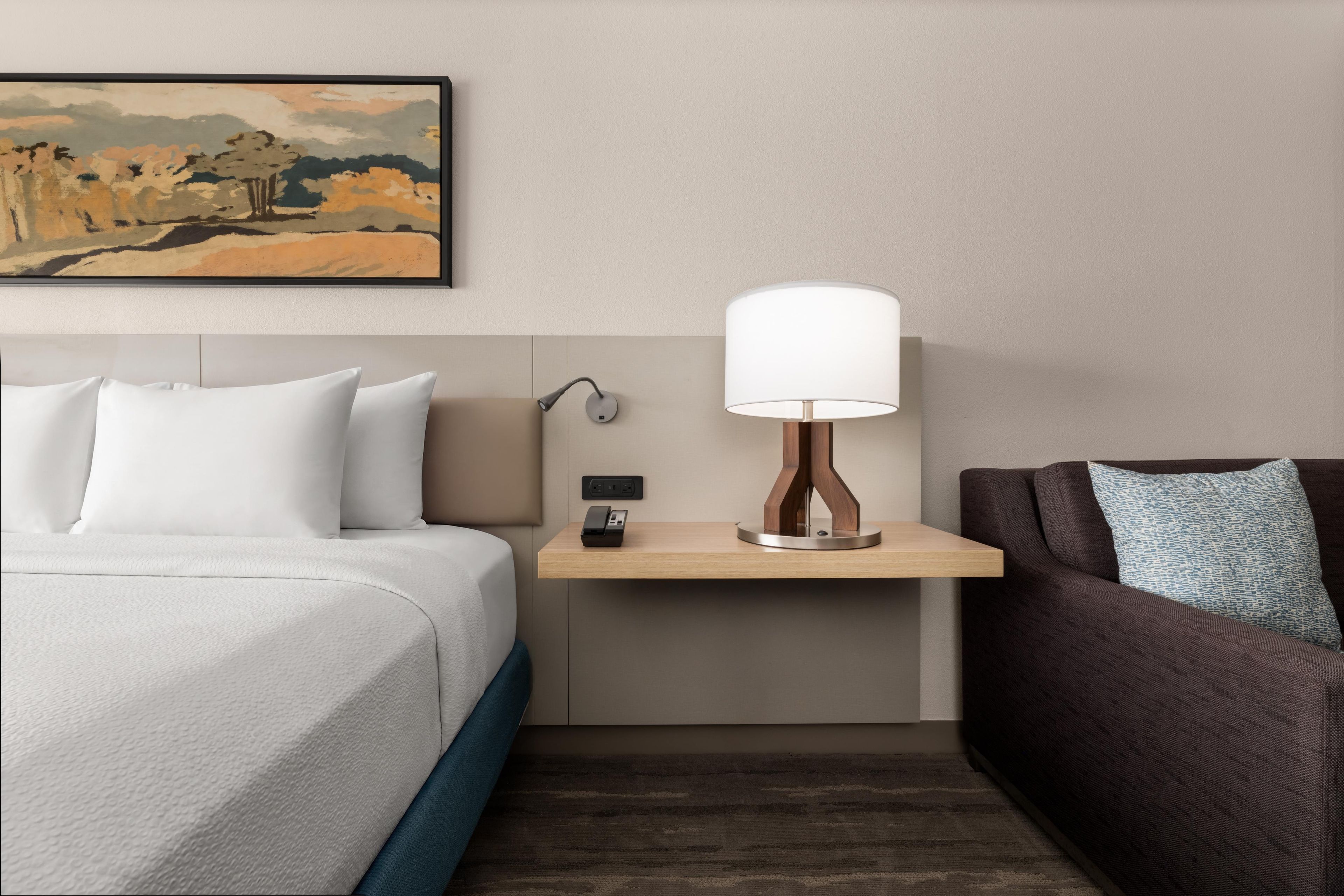 Habitación de hotel con cama, mesita de noche con lámpara, teléfono y un cuadro en la pared.