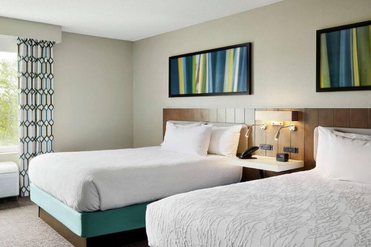 A hotel room with two beds, a window with a geometric curtain, and a wall-mounted artwork.