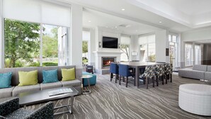 A spacious living area with a fireplace, a dining table with chairs, a sofa with cushions, and large windows offering a view of the outdoors. - Hilton Garden Inn Cincinnati Northeast (Loveland)