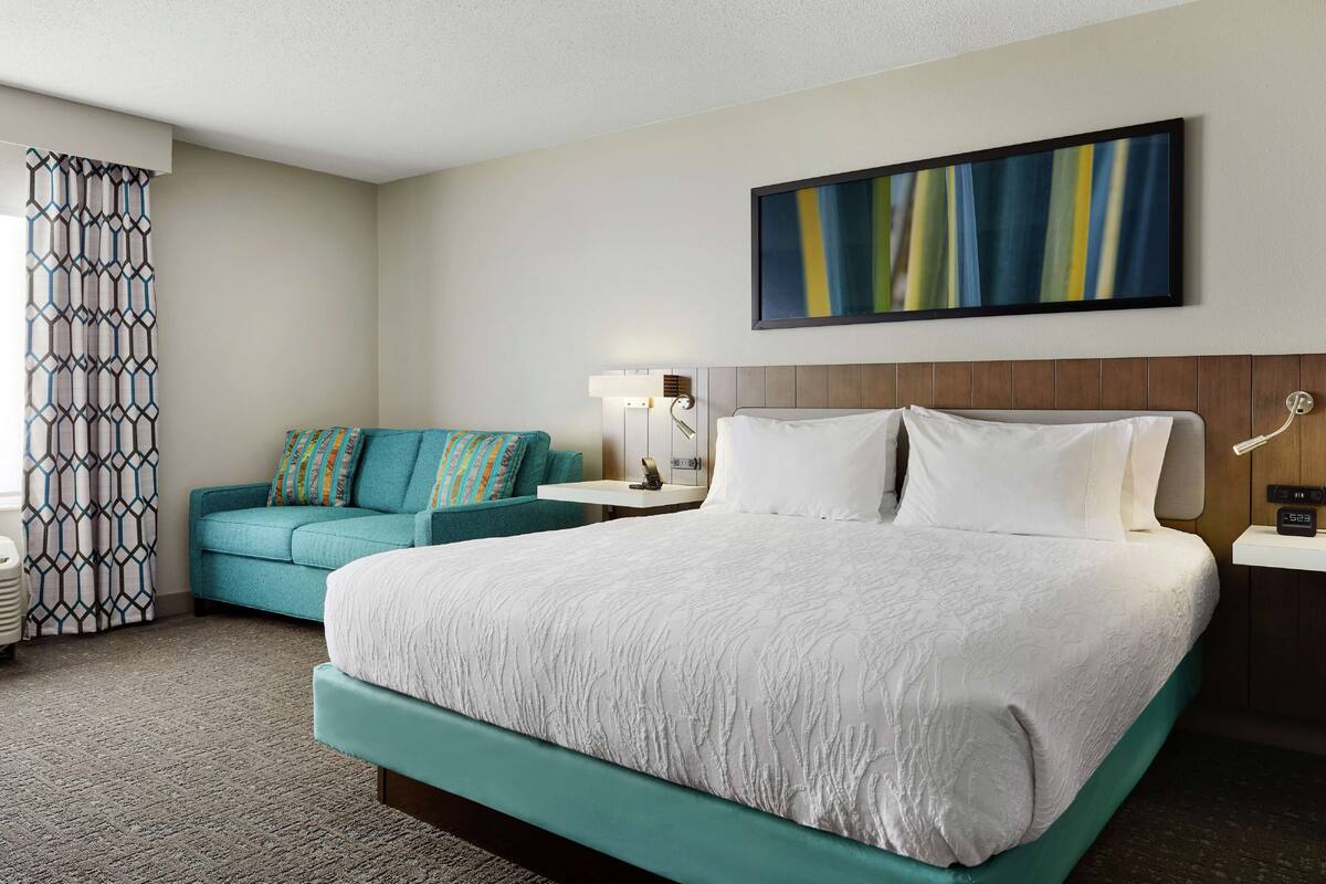 A hotel room with a large bed, a sofa, a nightstand, a lamp, and a painting on the wall.