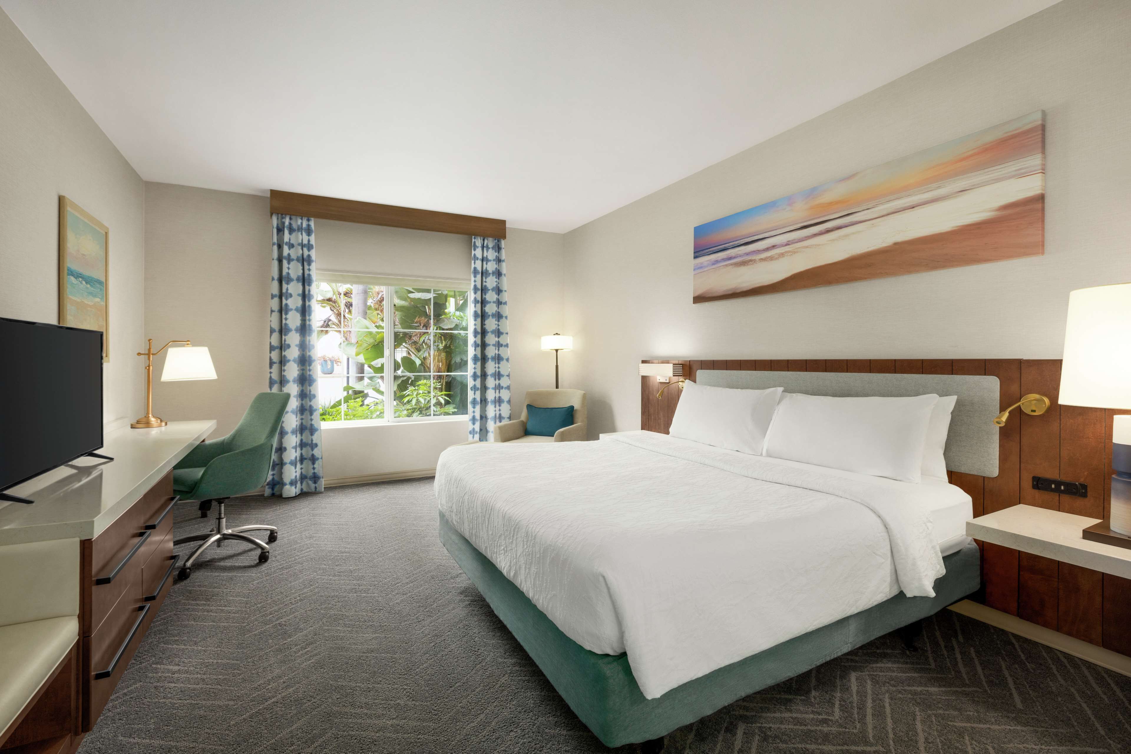 A hotel room with a bed, desk, chair, TV, and a window with curtains.
