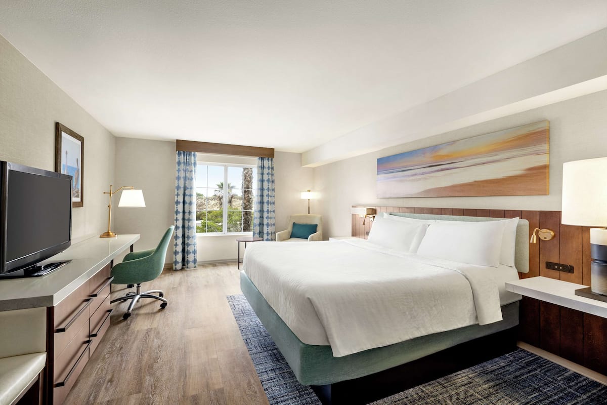 A hotel room with a bed, a desk with a TV, a chair, and a window with curtains.