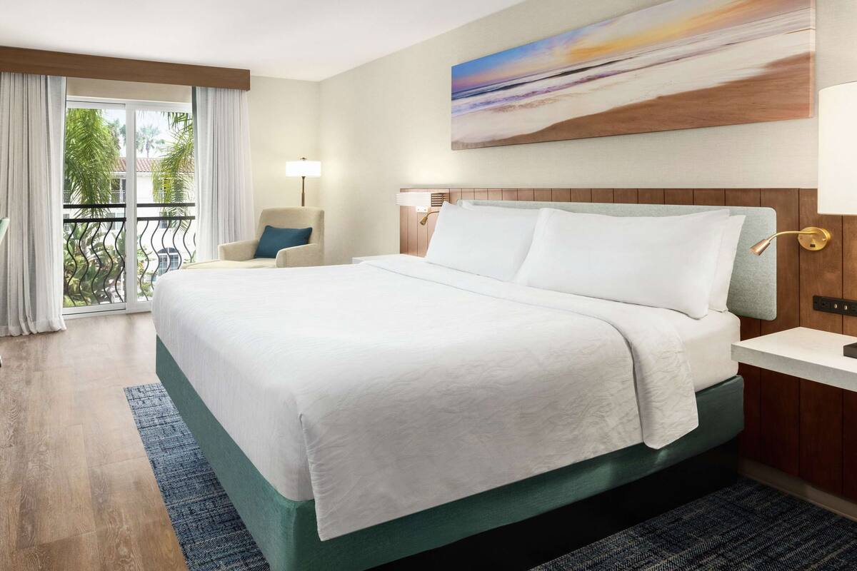 A hotel room with a large bed, a chair, a painting, and a view of the outdoors.