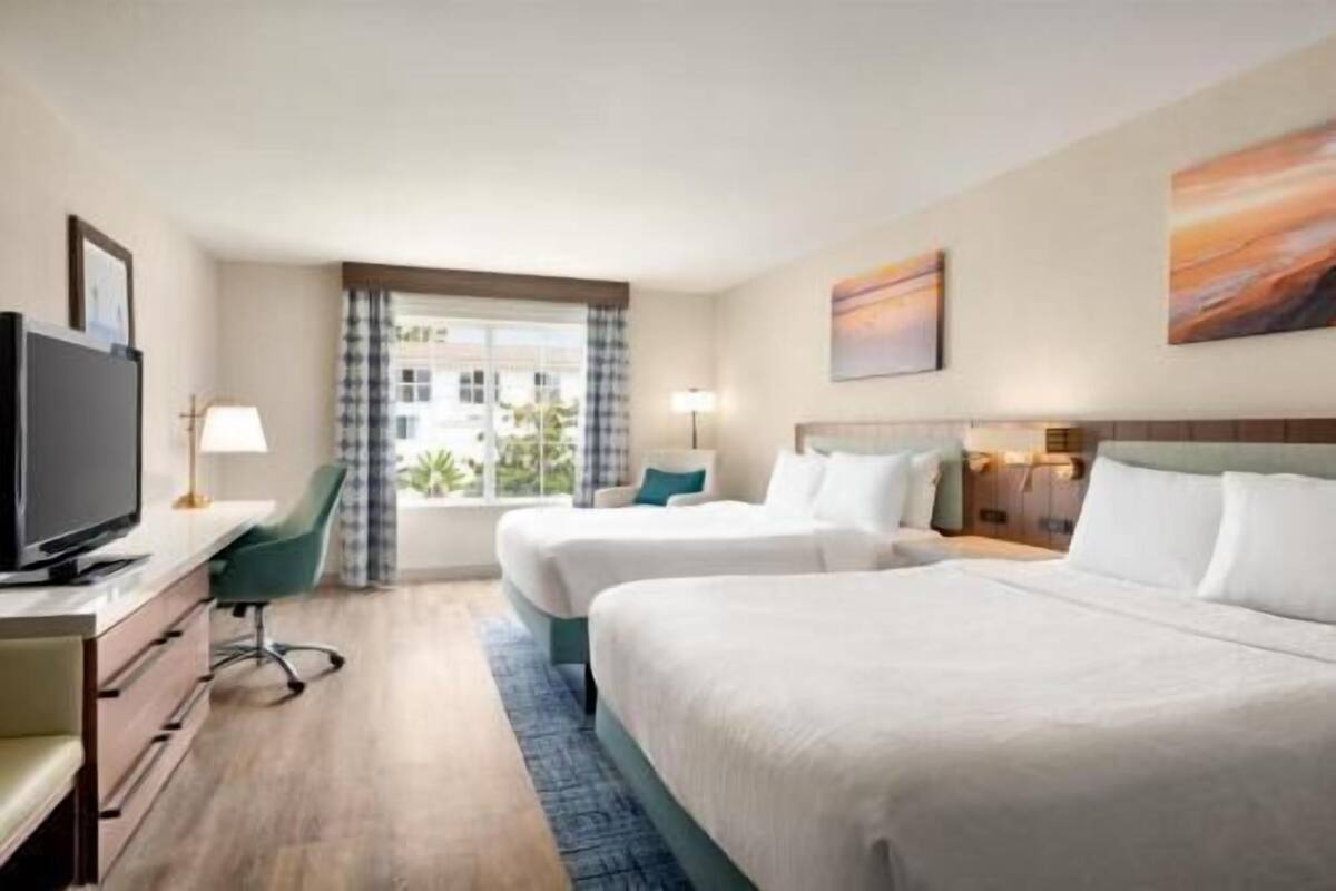 A hotel room with two beds, a desk with a TV, a window with curtains, and a painting on the wall.