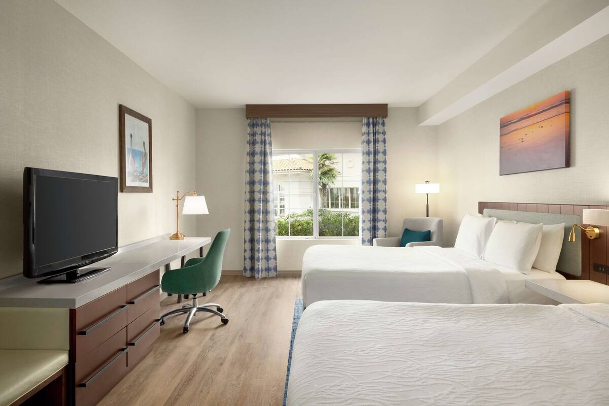 A hotel room with a TV, desk, two beds, a chair, and a window with curtains.