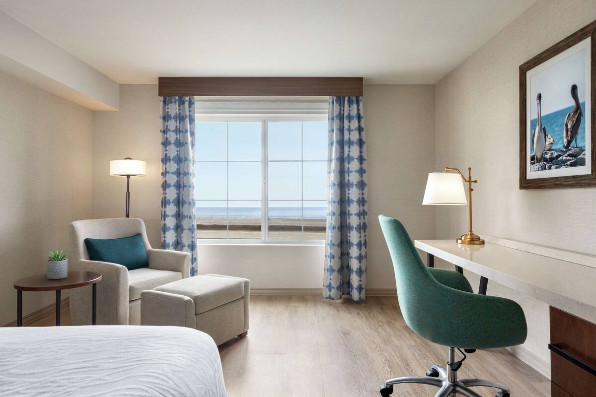 A hotel room with a bed, a desk, a chair, a small table with a lamp, and a window with curtains.