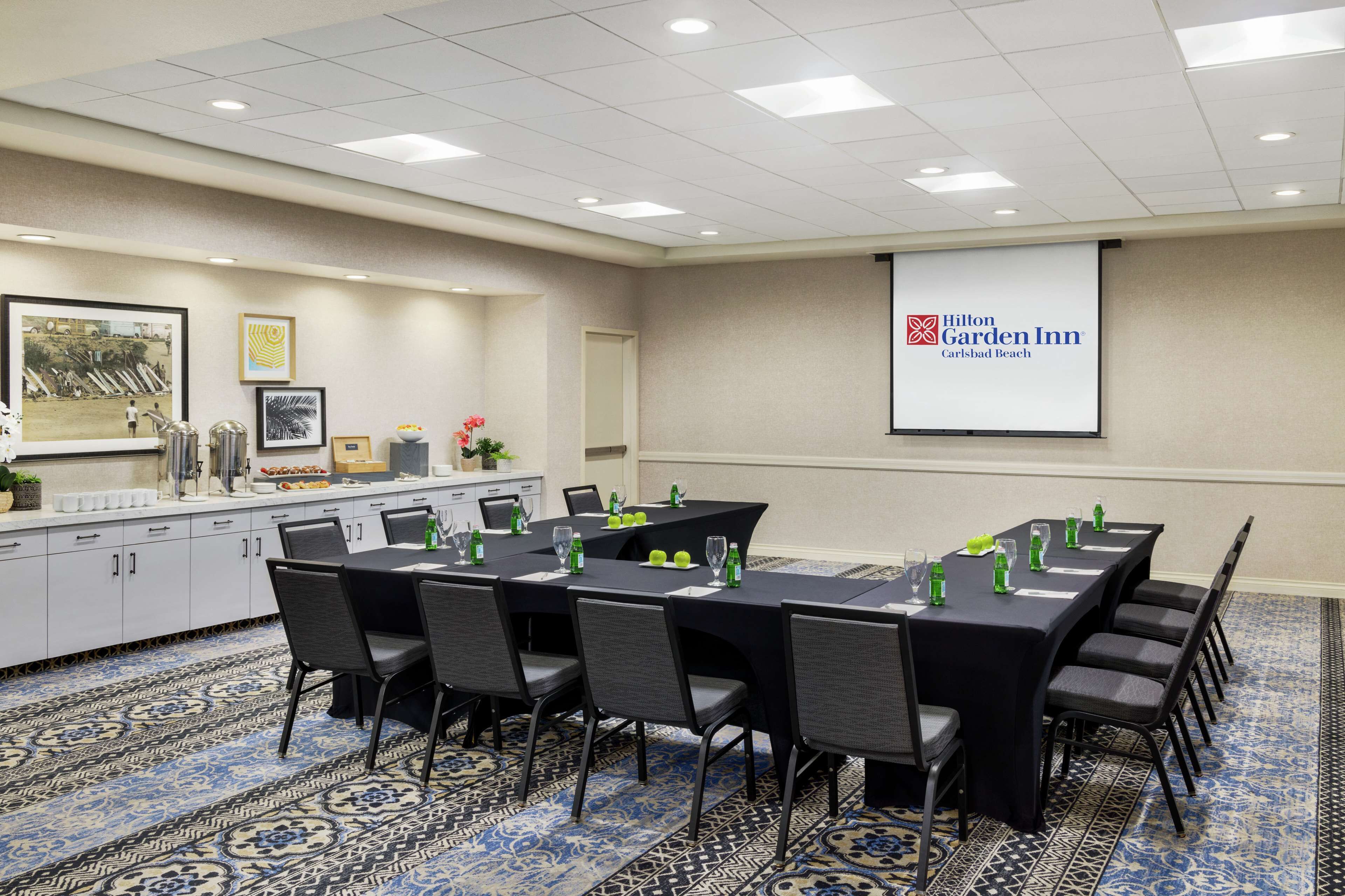 A conference room with a large screen displaying the Hilton Garden Inn logo, a long table with chairs, and a kitchenette area.