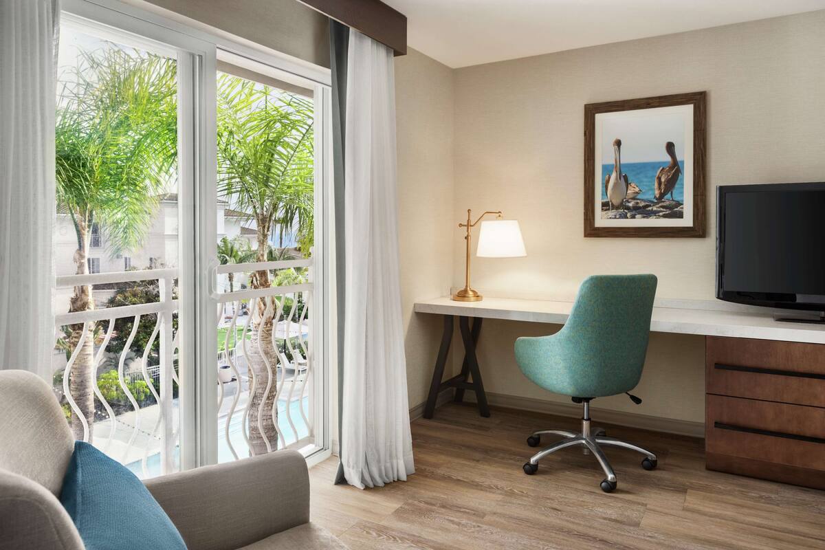 A hotel room with a large window, a desk with a lamp, a television, and a comfortable seating area.