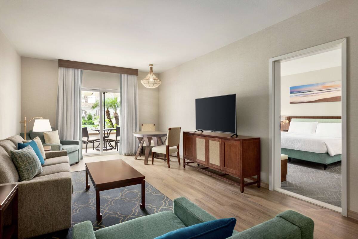 A hotel room with a sofa set, a TV on a wooden cabinet, a dining table with chairs, and a large window with curtains.