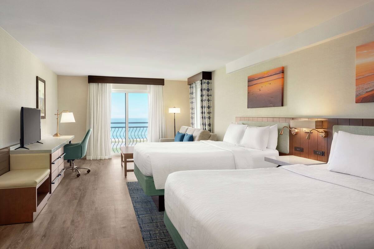 A hotel room with two beds, a desk, a TV, and a view of the ocean.