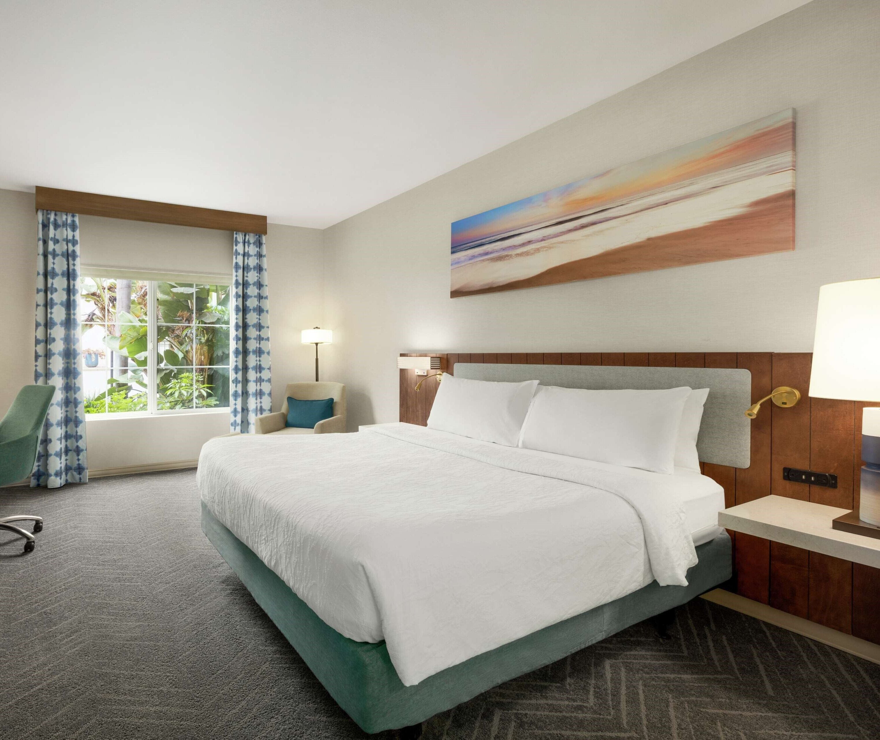 A hotel room with a large bed, a desk, a chair, a window with curtains, and a painting on the wall.