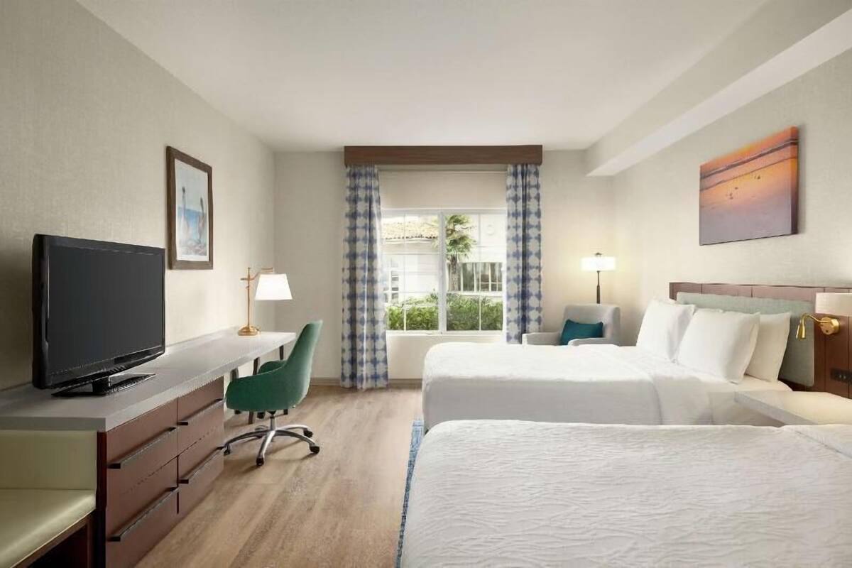 A hotel room with a bed, a desk with a television, a chair, a lamp, and a window with curtains.