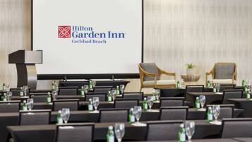 A conference room at Hilton Garden Inn with a large screen displaying the hotel's logo, rows of chairs, and tables set with water bottles and glasses.