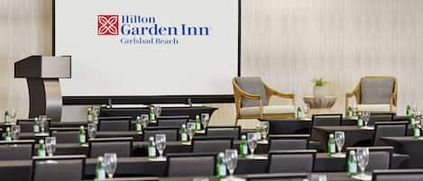 A conference room at Hilton Garden Inn with a large screen displaying the hotel's logo, rows of chairs, and tables set with water bottles and glasses.