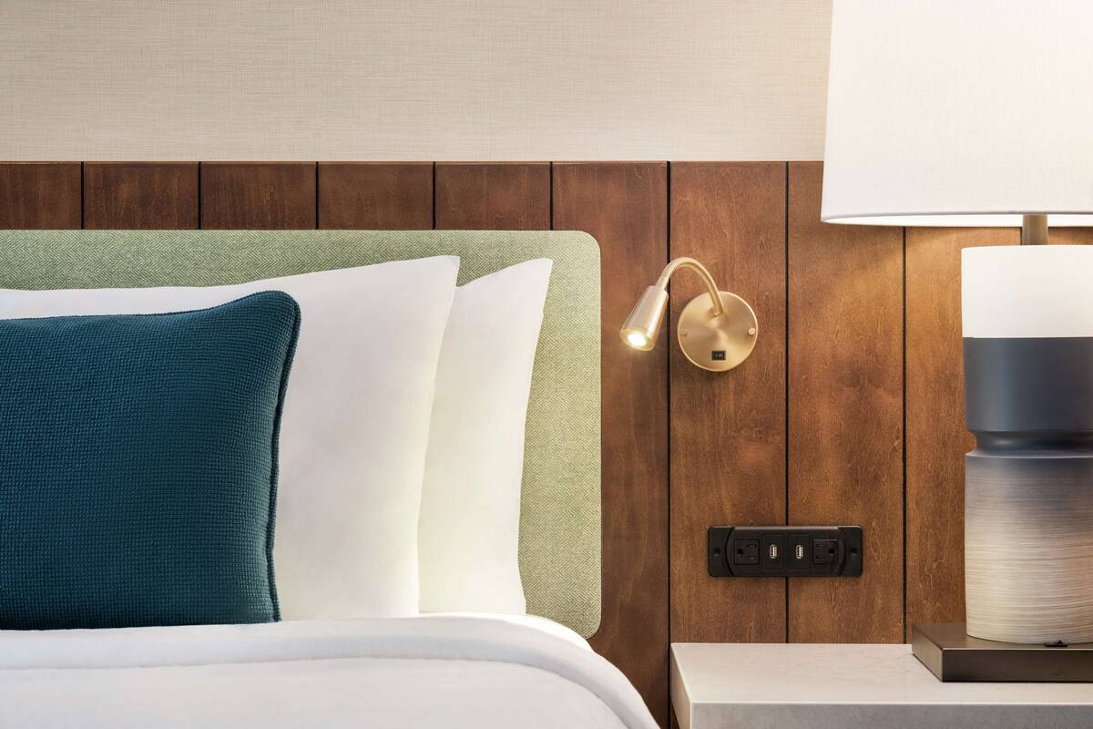 A neatly made bed with white and teal pillows, a green headboard, a wall-mounted lamp, and a power outlet on a wooden panel wall.