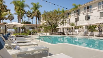 A pool area with lounge chairs and umbrellas, surrounded by palm trees and a building with balconies.