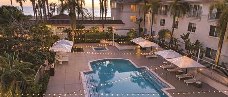 A pool area with lounge chairs and umbrellas, surrounded by palm trees and a building with balconies.